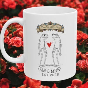 May include: White ceramic mug featuring a whimsical design of two cartoon frogs facing each other with a red heart between them. The mug has the text "Thanks, Algorithm" above the frogs and "EVAN & BENNY EST.2025" below. The mug has a curved handle.