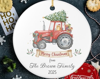 May include: A white ceramic Christmas ornament featuring a red tractor carrying a Christmas tree and gifts. The ornament includes the text "Merry Christmas from The Brown Family 2025". A small cat is in the tractor.
