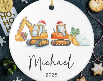 Pode incluir: Enfeite de Natal de cerâmica branca com dois veículos de construção de desenho animado, um amarelo e outro laranja, usando gorros de Papai Noel. O enfeite tem o nome "Michael" e o ano "2025" impressos.
