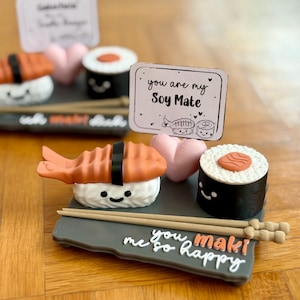 Könnte beinhalten: Dekoratives Sushi-Set mit einem lächelnden Shrimp-Nigiri, einer Maki-Rolle und einem rosa Herz. Das Set enthält ein graues Tablett mit dem Text "you maki me so happy" und eine Karte mit der Aufschrift "you are my Soy Mate".