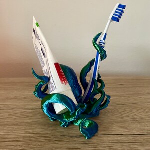 May include: A teal and green 3D printed octopus holding a tube of toothpaste and a blue toothbrush. The octopus is designed to hold both items upright.