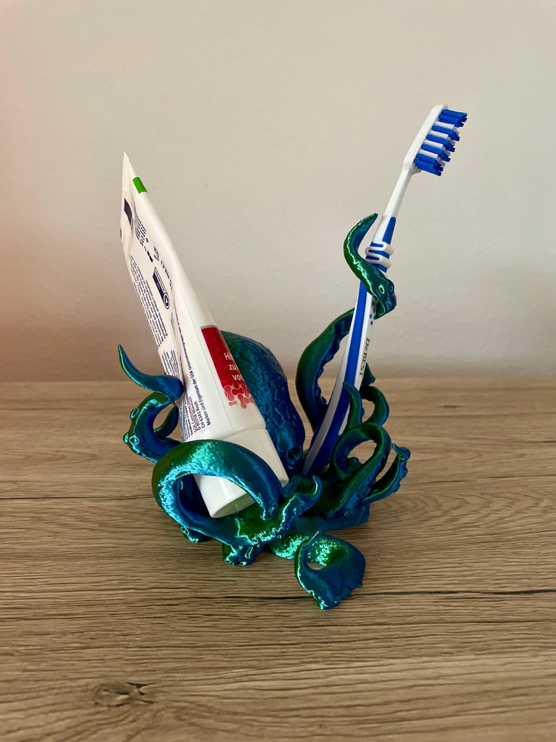 May include: A teal and green 3D printed octopus holding a tube of toothpaste and a blue toothbrush. The octopus is designed to hold both items upright.