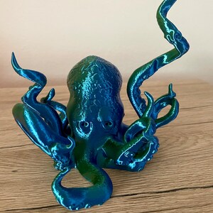 May include: A 3D printed octopus figurine with a blue and green iridescent finish. The octopus has eight tentacles and is posed with its tentacles raised.