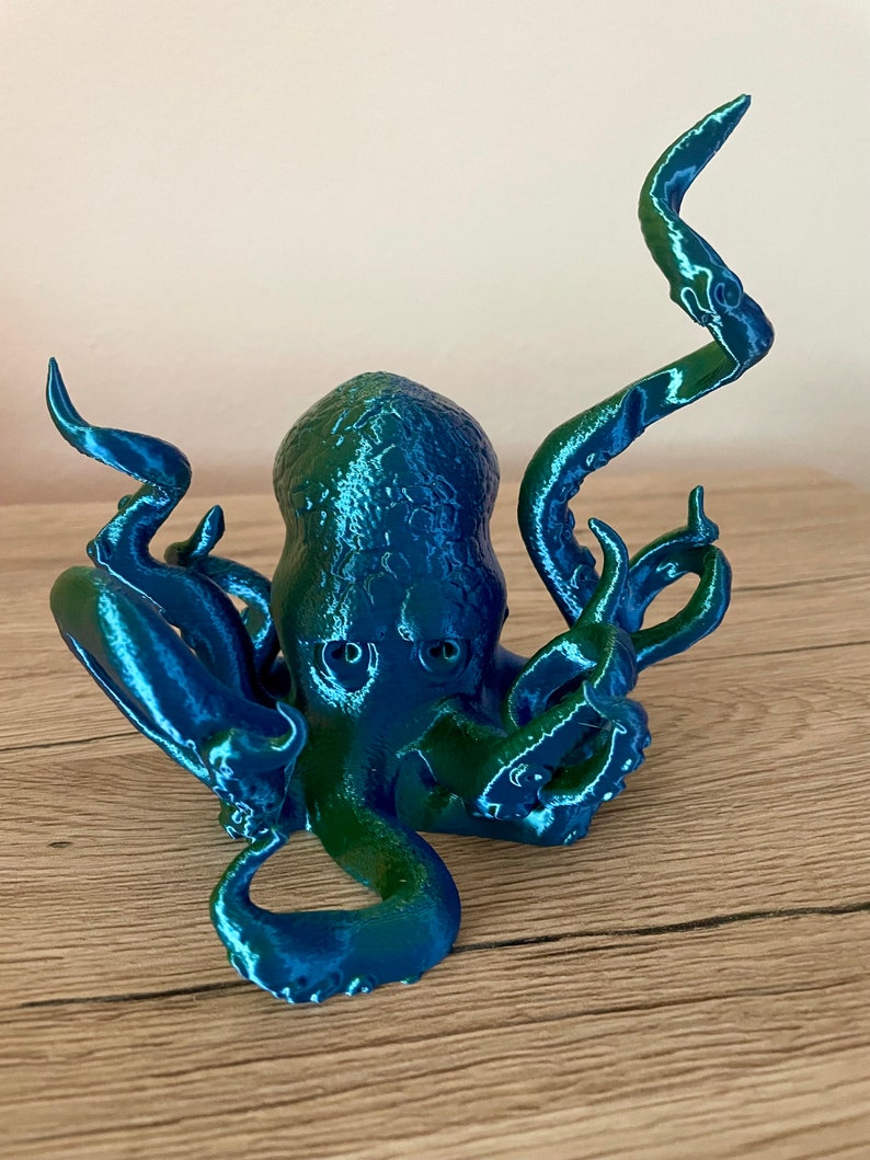 May include: A 3D printed octopus figurine with a blue and green iridescent finish. The octopus has eight tentacles and is posed with its tentacles raised.