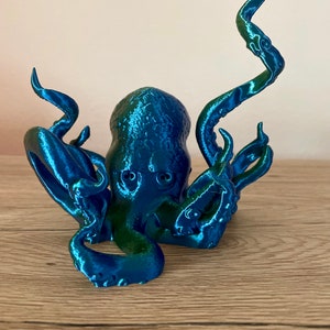 May include: A 3D printed octopus figurine with a blue and green iridescent finish. The octopus has eight tentacles and is posed in a sitting position.