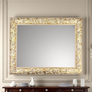 Peut inclure: Grand miroir rectangulaire avec un cadre orné. Le cadre présente une base blanche avec des accents de feuilles d'or, créant un aspect texturé et élégant. Le miroir est fixé sur un mur neutre au-dessus d'un meuble en bois foncé.