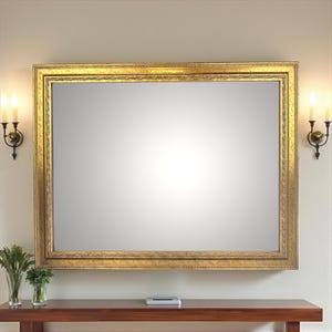 Może przedstawiać: Duże prostokątne lustro ze złotą, ozdobną ramą wisi nad drewnianym stołem konsolowym. Lustro odbija pokój. Dwa kinkiety z zapalonymi świecami flankują lustro. Wazon z zielenią stoi na stole.