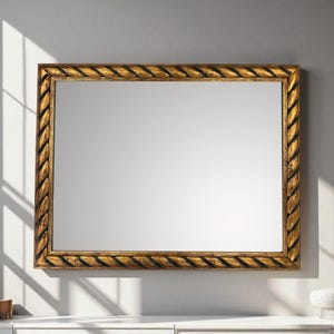 Puede incluir: Espejo rectangular con marco dorado con diseño de cuerda. El espejo está montado sobre una superficie blanca contra una pared gris. El marco tiene un acabado envejecido, lo que añade una estética vintage.