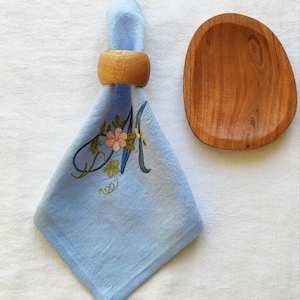 Può includere: Un tovagliolo di lino azzurro con un ricamo floreale e la lettera "A" è tenuto da un anello portatovagliolo in legno. A destra si trova una ciotola di legno di forma ovale.