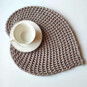 May include: A beige, hand-knitted placemat with a leaf-like shape. A white teacup and saucer with a floral pattern sit on the placemat. The placemat is made of thick, textured yarn, and the teacup and saucer are made of porcelain.