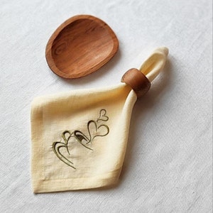Può includere: Un piatto di legno e un tovagliolo giallo piegato con cuori verdi ricamati. Un anello per tovagliolo marrone tiene il tovagliolo. Gli oggetti sono disposti su una superficie bianca, suggerendo un tema di sala da pranzo o tavola.