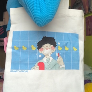 May include: White canvas tote bag with a blue tiled bathroom scene featuring a cartoon character brushing their teeth with a red toothbrush. The character has a white towel around their head and is surrounded by yellow rubber duckies. The text "BRANDYYOMOSS" is printed at the bottom of the scene.
