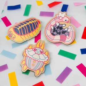 Puede incluir: Tres pines de madera con ilustraciones coloridas. Uno representa un insecto segmentado amarillo y azul. Otro muestra una criatura rosa y morada con un lazo. El tercero presenta un diseño de cupcake azul y rosa.