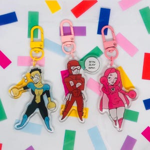 Op de afbeelding: Drie acryl sleutelhangers met cartoon superhelden ontwerpen. De sleutelhangers hebben roze, gele en gouden sluitingen. Eén superheld draagt een rood pak, een ander een zwart en blauw pak en de derde een roze pak. Eén sleutelhanger heeft een tekstballon met "hit the SLAY button."