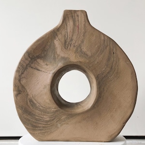 Op de afbeelding: Een lichtbruine keramische vaas met een unieke, abstracte vorm. De vaas heeft een grote, ronde opening in het midden.
