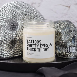 May include: A white candle with a black label that reads "Tattoos Pretty Eyes & Thick Thighs". The candle is sitting on a black tray with two silver skull figurines.