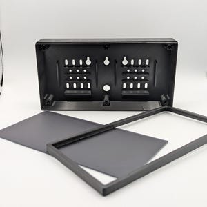 May include: A black rectangular plastic housing with a grid of mounting holes and keyhole slots on the back panel, accompanied by a separate thin black rectangular frame and a flat black rectangular insert piece for assembly.