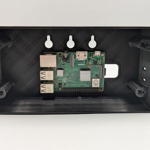 May include: A black rectangular 3D-printed plastic enclosure featuring three keyhole mounting slots on the top edge and four corner screw pillars. A green Raspberry Pi circuit board with silver heat sinks and various ports is mounted inside.