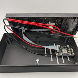 Pode incluir: Caixa de dispositivo eletr&ocirc;nico preta aberta, revelando uma placa de circuito e fia&ccedil;&atilde;o. Fios vermelhos e pretos est&atilde;o conectados &agrave; se&ccedil;&atilde;o superior, enquanto uma pequena placa de circuito est&aacute; dentro. A caixa est&aacute; aberta.