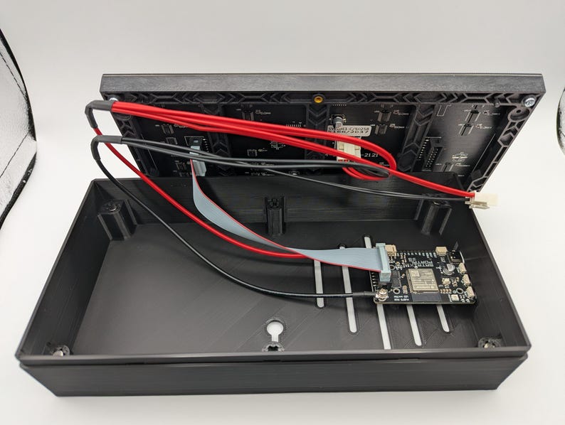 Pode incluir: Caixa de dispositivo eletr&ocirc;nico preta aberta, revelando uma placa de circuito e fia&ccedil;&atilde;o. Fios vermelhos e pretos est&atilde;o conectados &agrave; se&ccedil;&atilde;o superior, enquanto uma pequena placa de circuito est&aacute; dentro. A caixa est&aacute; aberta.