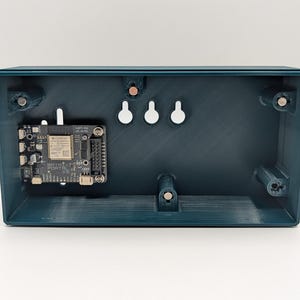 Pode incluir: Uma caixa retangular aberta, de cor verde-azulado, revelando uma placa de circuito e hardware de montagem. A placa de circuito preta tem v&aacute;rios componentes. Tr&ecirc;s pontos de montagem brancos e circulares est&atilde;o na parede traseira.