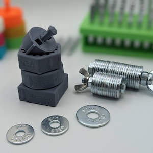 May include: A set of three gray 3D printed Ethereum cryptocurrency tokens, a spinning top, and three silver washers with the words "CYPRO", "SECO", and "STACKER" engraved on them. The tokens are stacked on top of each other, with the top token being the spinning top. The washers are scattered around the tokens.
