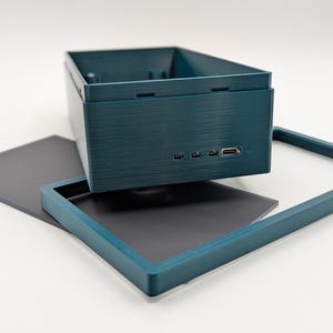 Pode incluir: Uma caixa retangular azul-petr&oacute;leo com superf&iacute;cie texturizada, apresentando v&aacute;rias portas pequenas e uma porta USB. Uma moldura azul-petr&oacute;leo correspondente e uma folha retangular cinza escuro tamb&eacute;m est&atilde;o presentes, tudo sobre um fundo branco.