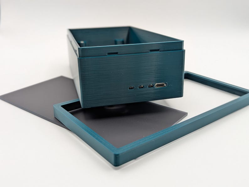 Pode incluir: Uma caixa retangular azul-petr&oacute;leo com superf&iacute;cie texturizada, apresentando v&aacute;rias portas pequenas e uma porta USB. Uma moldura azul-petr&oacute;leo correspondente e uma folha retangular cinza escuro tamb&eacute;m est&atilde;o presentes, tudo sobre um fundo branco.