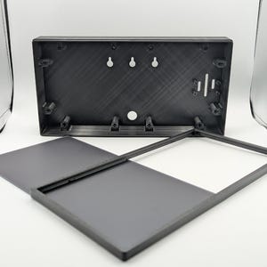 May include: A black rectangular plastic enclosure with a separate flat cover. The main housing features internal mounting pillars, three keyhole slots for wall mounting, and a circular opening. The cover is a thin, smooth, rectangular panel.