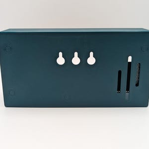 Pode incluir: Uma caixa de armazenamento retangular montada na parede, na cor verde-azulado. A parte traseira da caixa apresenta tr&ecirc;s ranhuras em forma de fechadura para pendurar, juntamente com duas ranhuras verticais e uma pequena abertura ranhurada. A caixa tem um acabamento liso e mate.