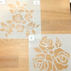 May include: Two floral stencils on a wooden surface. One stencil features a detailed rose bouquet design, while the other showcases a simpler rose arrangement. Each stencil is labeled with a letter or number in the corner.