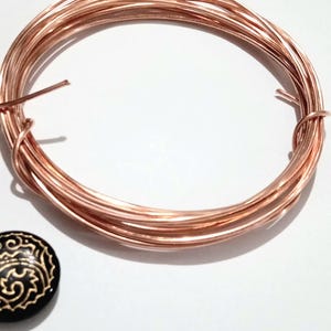 以下が含まれることがあります： 円形のループ状に配置された光沢のある金属製の銅線コイルと、表面に複雑な金色の渦巻き模様が刻まれた小さな丸い黒いボタン。銅線は滑らかで反射する仕上げが施されており、ボタンの隣に配置されています。