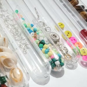 May include: Assortment of clear glass test tubes filled with various colorful beads, buttons, seashells, and other small objects. The test tubes are arranged in a row on a white surface.