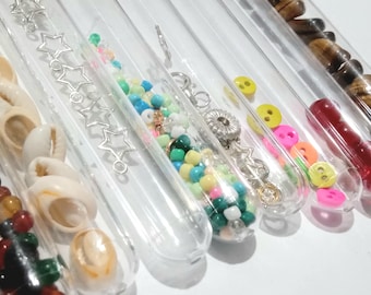 Op de afbeelding: Assortiment van heldere glazen reageerbuizen gevuld met verschillende gekleurde kralen, knopen, schelpen en andere kleine voorwerpen. De reageerbuizen zijn in een rij op een wit oppervlak gerangschikt.