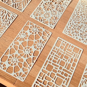 Könnte beinhalten: Eine Sammlung weißer, rechteckiger Schablonen mit verschiedenen ausgeschnittenen Mustern, darunter Blumen-, geometrische und abstrakte Designs. Die Schablonen sind auf einer Holzoberfläche angeordnet und zeigen ihre komplizierten Details und ihr Potenzial für Bastelprojekte.