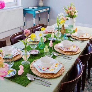 May include: An Easter table setting with floral arrangements, pastel tableware, and bunny decorations. The table features a green runner, woven placemats, square plates, and bowls. The scene is set for a festive celebration with a spring theme.