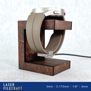 May include: A wooden watch stand with a brown finish, holding a silver smartwatch with a gray silicone band. The stand has a small cutout for the charging cable. The text "LASER FILECRAFT" and "3mm | 3.175mm | 1/8" | 4mm" is visible at the bottom of the image.