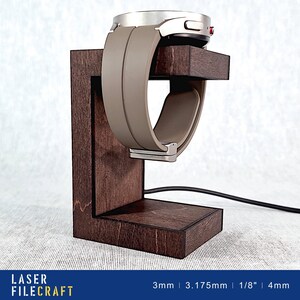 May include: A dark brown wooden watch stand with a silver watch with a gray band resting on it. The stand has a slot for charging the watch. The text "LASER FILECRAFT" is printed on the bottom of the image.