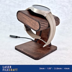 Pode incluir: Um suporte de carregamento de madeira para um smartwatch. O suporte é feito de madeira marrom escura e tem uma ranhura para o relógio se sentar. O relógio é um smartwatch prateado com uma banda preta. O suporte tem 3 mm de espessura e uma ranhura de 3,8 mm para o relógio.