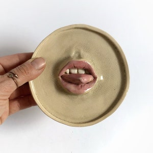 Pode incluir: Um prato de cer&acirc;mica com uma imagem esculpida em relevo de uma boca com a l&iacute;ngua de fora. O prato &eacute; de cor bege claro com uma superf&iacute;cie ligeiramente texturizada.