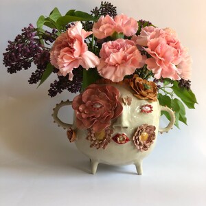 Könnte beinhalten: Eine weiße Keramikvase mit einem Gesicht, das auf der Vorderseite modelliert ist. Die Vase ist mit rosa und orangefarbenen Blumen verziert und hat drei Beine. Die Vase ist mit rosa Nelken und lila Flieder gefüllt.