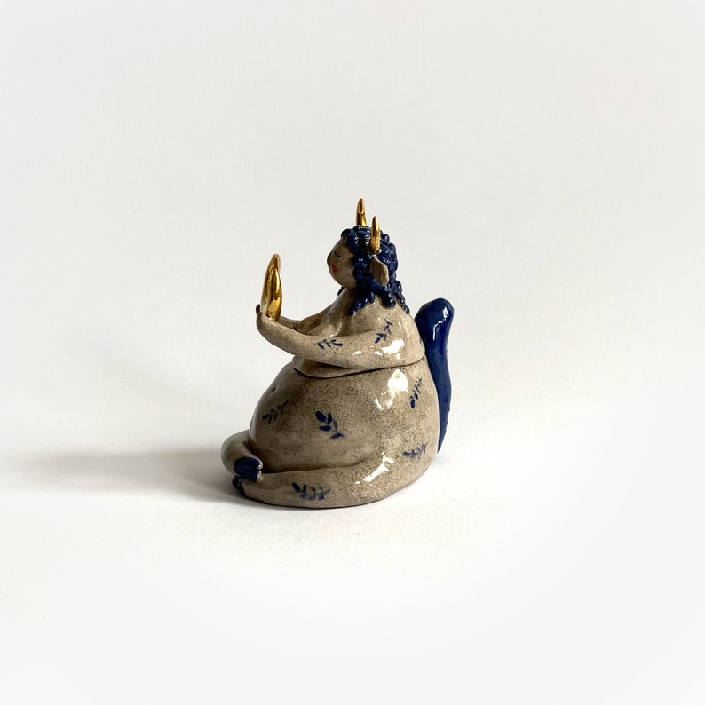 May include: A ceramic sculpture of a seated figure with blue hair and a blue tail. The figure holds a gold-colored object and is adorned with gold horns. The figure's body is beige with blue floral patterns. The sculpture is on a white background.