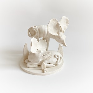Puede incluir: Escultura de cerámica blanca de dos animales estilizados con orejas grandes, uno de pie y el otro acostado, rodeado de hojas. La escultura está sobre una base circular blanca.