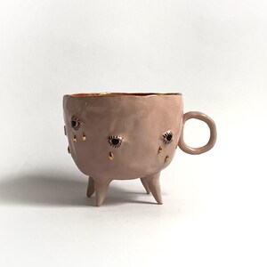 May include: A pink ceramic mug with three legs and gold accents. The mug has black eyes and gold teardrop designs.