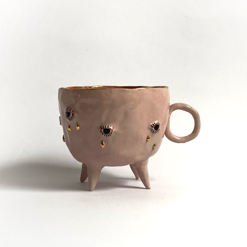 May include: A pink ceramic mug with three legs and gold accents. The mug has black eyes and gold teardrop designs.