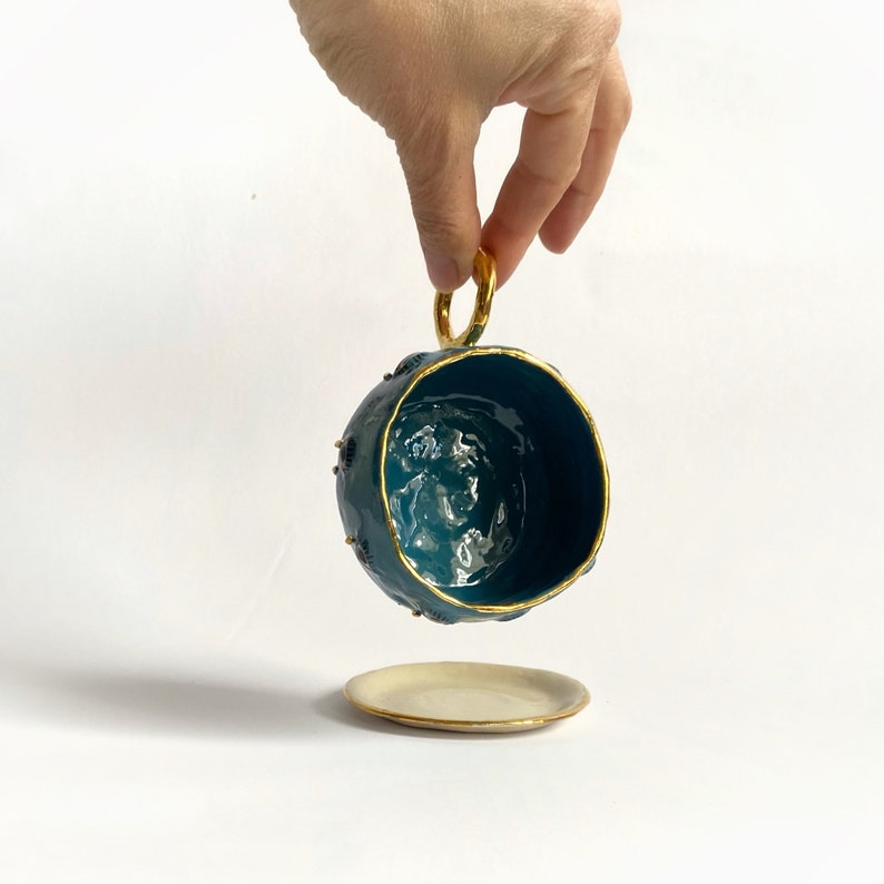 K&ouml;nnte beinhalten: Eine teeblau gef&auml;rbte Keramiktasse mit goldfarbenem Rand und Henkel. Die Tasse ist mit kleinen goldenen Punkten verziert und steht auf einer wei&szlig;en Untertasse mit goldfarbenem Rand.