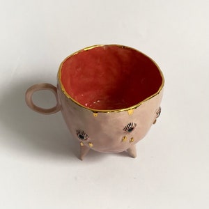 May include: A pink ceramic mug with three legs and gold accents. The mug has three eyes with black pupils and gold eyelashes. The inside of the mug is red.