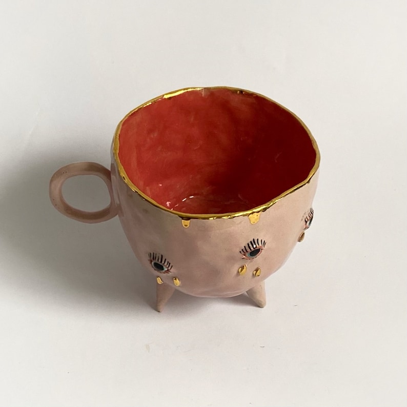 May include: A pink ceramic mug with three legs and gold accents. The mug has three eyes with black pupils and gold eyelashes. The inside of the mug is red.