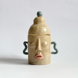 May include: A ceramic jar with a lid shaped like a head. The jar is beige with a crackled glaze and has a gold rim. The lid has a blue knob and the jar has two blue handles. The face on the jar has closed eyes and a pink mouth.