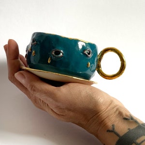 K&ouml;nnte beinhalten: Eine teeblaue Keramik-Teetasse mit goldfarbenem Rand und Henkel. Die Tasse hat drei bemalte Augen mit goldenen Tr&auml;nentropfen, die von jedem Auge tropfen.